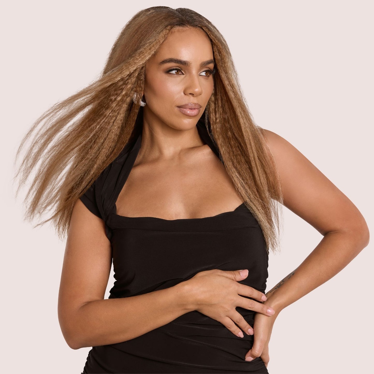 A confident woman with long, flowing kinky straight hair extensions in a golden brown shade. Her voluminous, blown-out strands move effortlessly as she poses elegantly in a sleek black dress. These premier textured hair extensions provide a seamless blend, adding fullness, length, and a natural-looking finish, perfect for versatile styling and a sophisticated appearance.