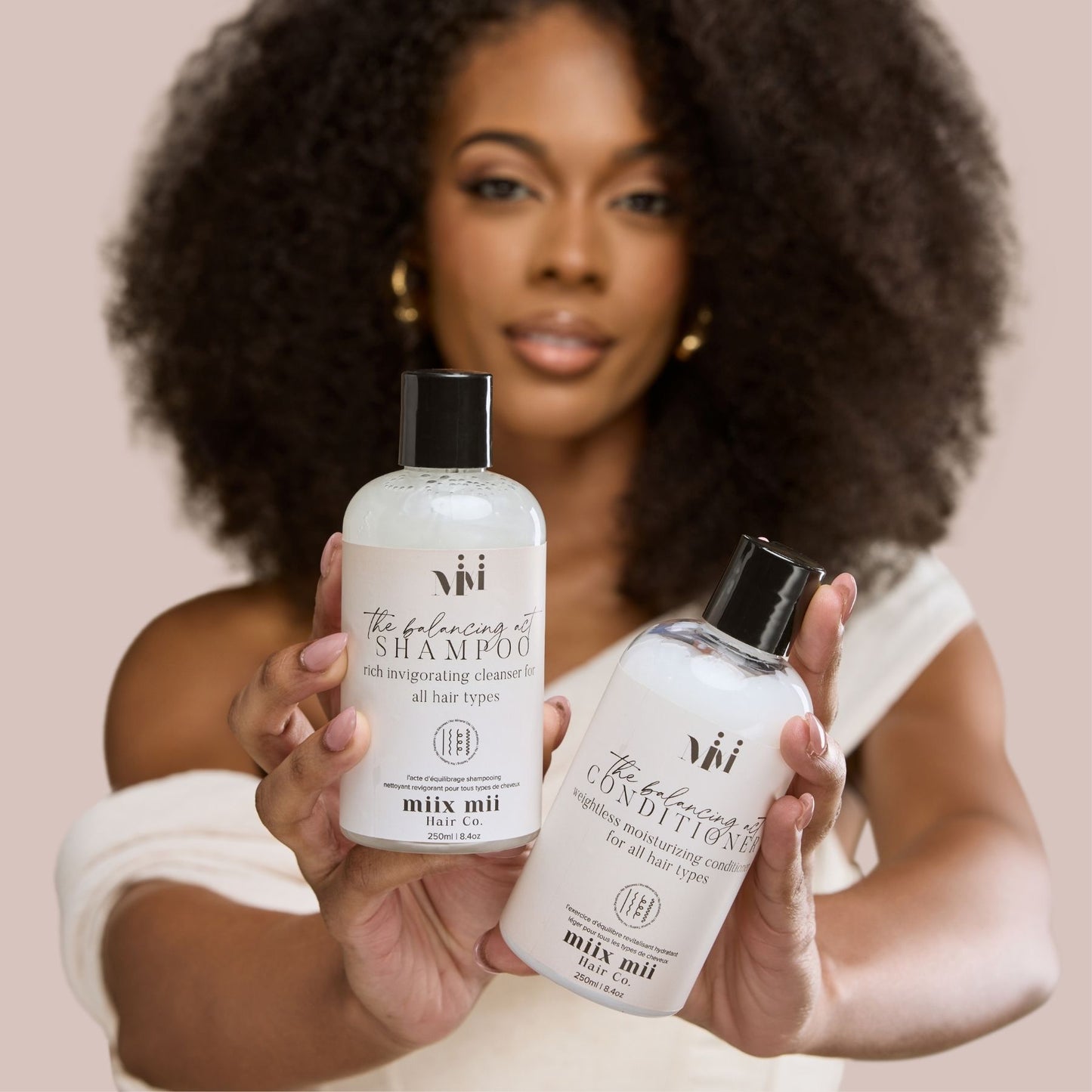 A confident Black woman with voluminous, natural-textured luxury hair extensions holds up two premium hair care products—shampoo and conditioner—from Miix Mii Hair Co. Her rich, bouncy curls exude health and shine, emphasizing the effectiveness of high-quality products for textured hair. The neutral-toned background and soft lighting highlight the elegance and sophistication of luxury hair care.