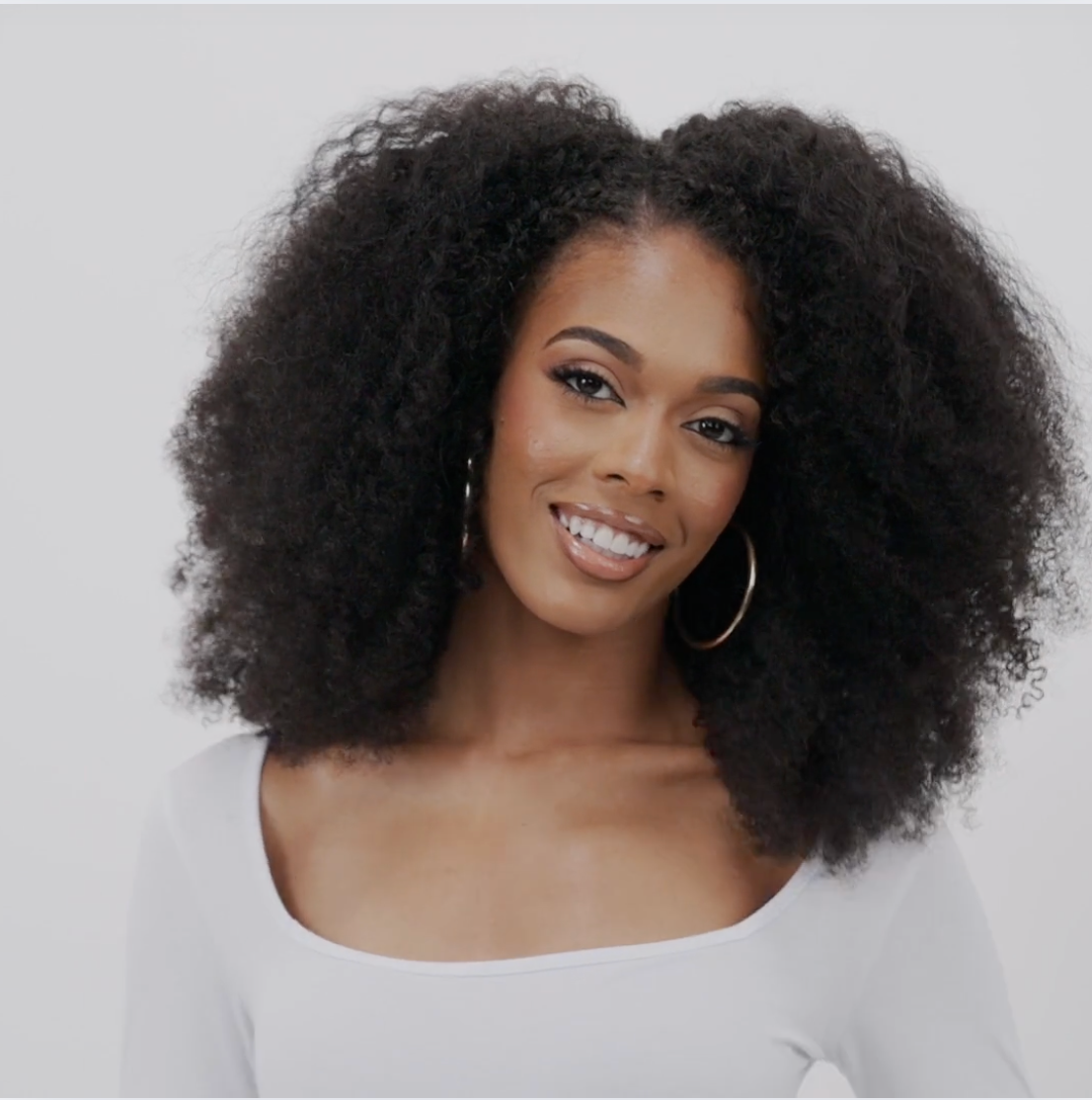A women wearing afro kinky curly hair extensions in a natural black shade, showcasing tight, voluminous curls with a natural texture. She moves her head from side to side to show movement and body in the hair extensions.